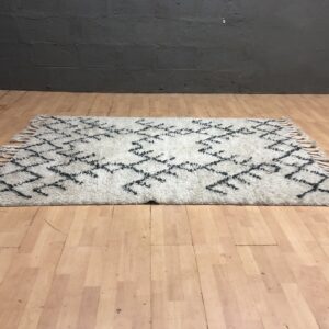 Cream shag pile area rug with dark geometric pattern and braided tassel fringe