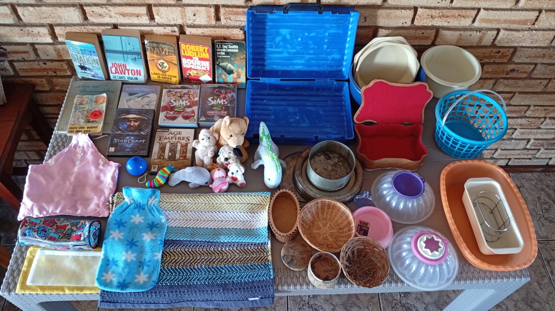 Mixed household lot ~30 items — books, PC game boxes (discs verified), plush toys, baking tins, baskets, mats, plasticware & blue case (clip missing)