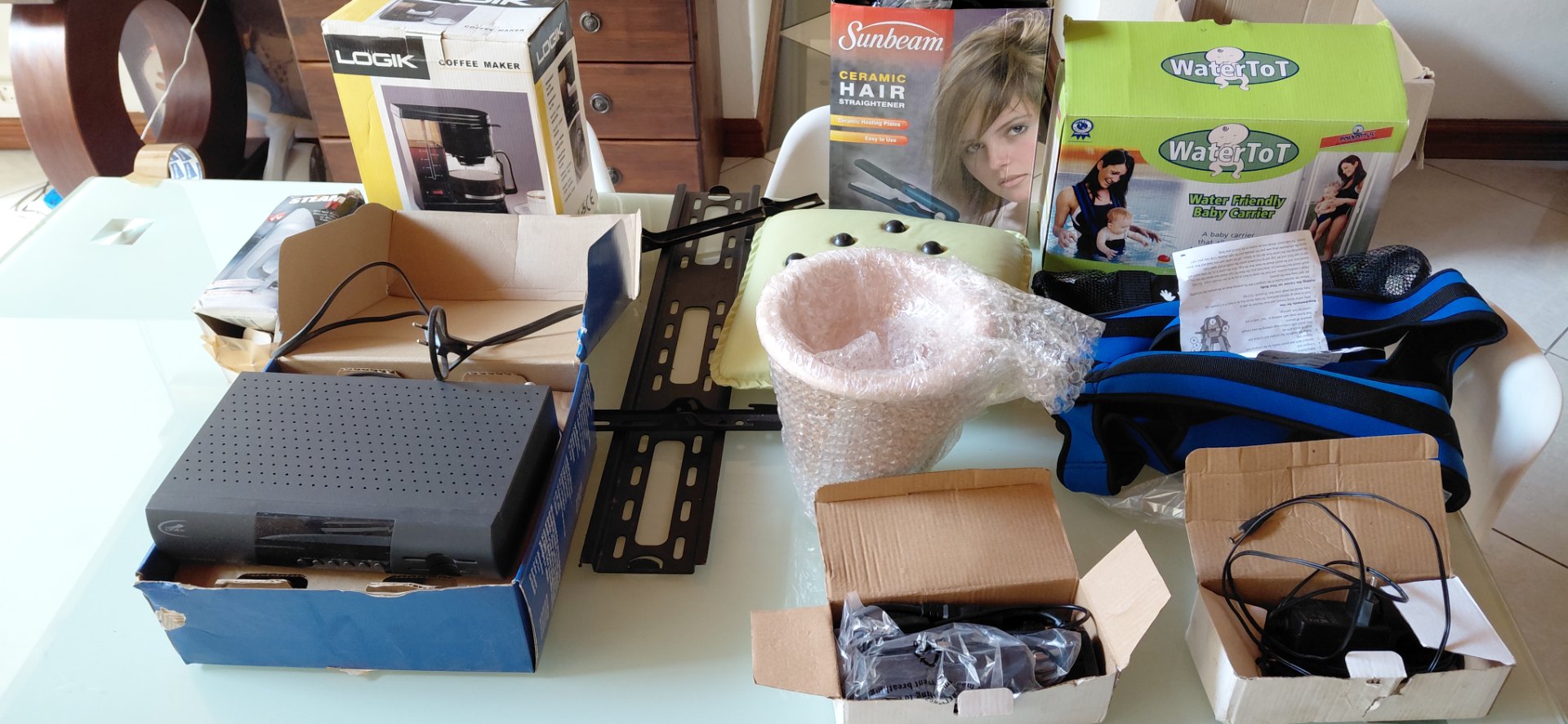 Lot 06 — Mixed household table lot: WaterToT baby carrier, boxed Logik coffee maker, Sunbeam hair straightener, set-top box, TV mount, cushion & accessories