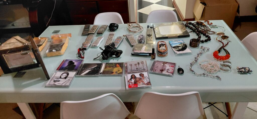 Lot 08 — Mixed table lot: packaged hair weaves, 2 wrapped wooden frames, costume jewellery, remotes, chargers, CDs & small accessories (Untested)