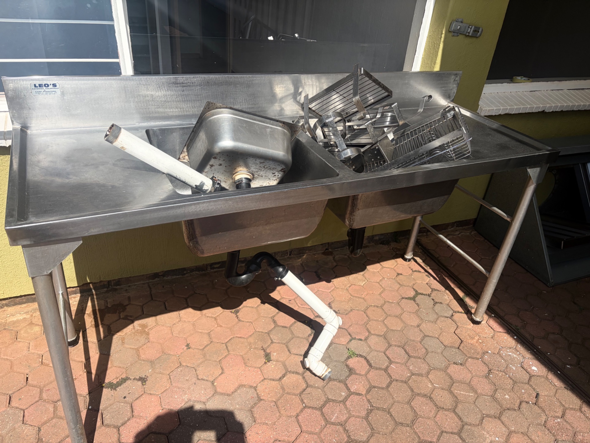 Commercial stainless steel double‑bowl sink with left drainboard on freestanding legs — used, accessories & loose plumbing