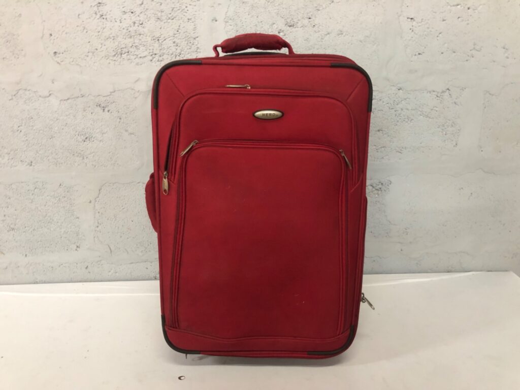 Red soft-sided wheeled suitcase — HERO badge, front pockets, wheels roll (62 x 24 x 40 cm)