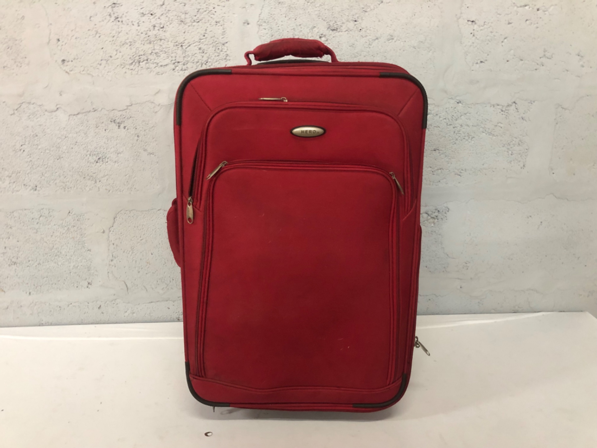 Red soft-sided wheeled suitcase — HERO badge, front pockets, wheels roll (62 x 24 x 40 cm)