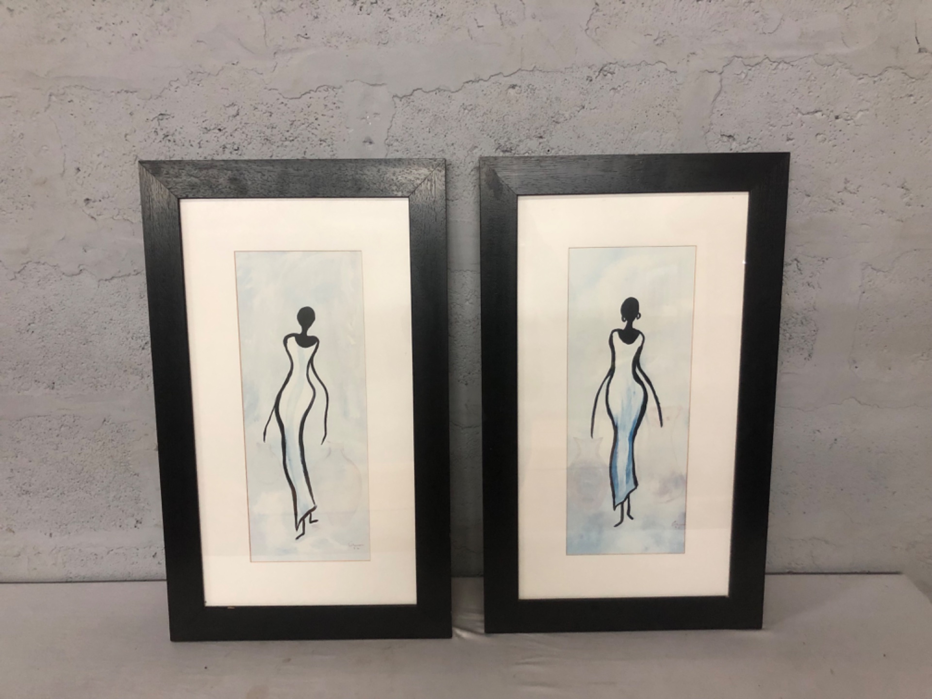 Pair of framed vertical artworks — stylised blue/black figures, black frames, glazed, 45 x 75 cm (each)