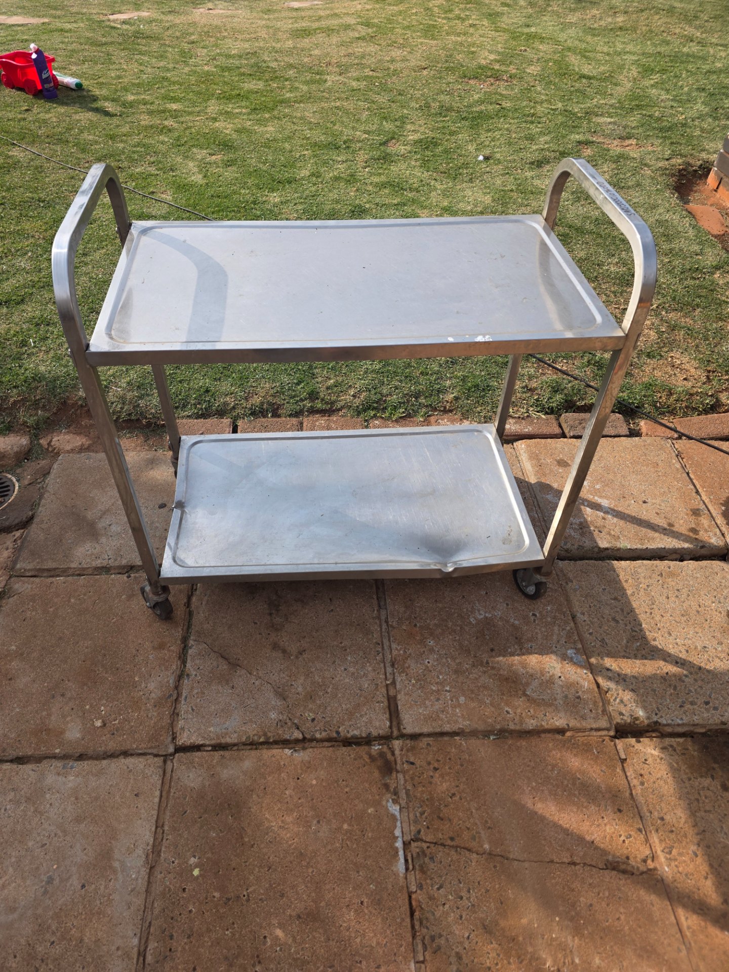 Two-shelf stainless-steel utility trolley on castor wheels — used, dented lower tray